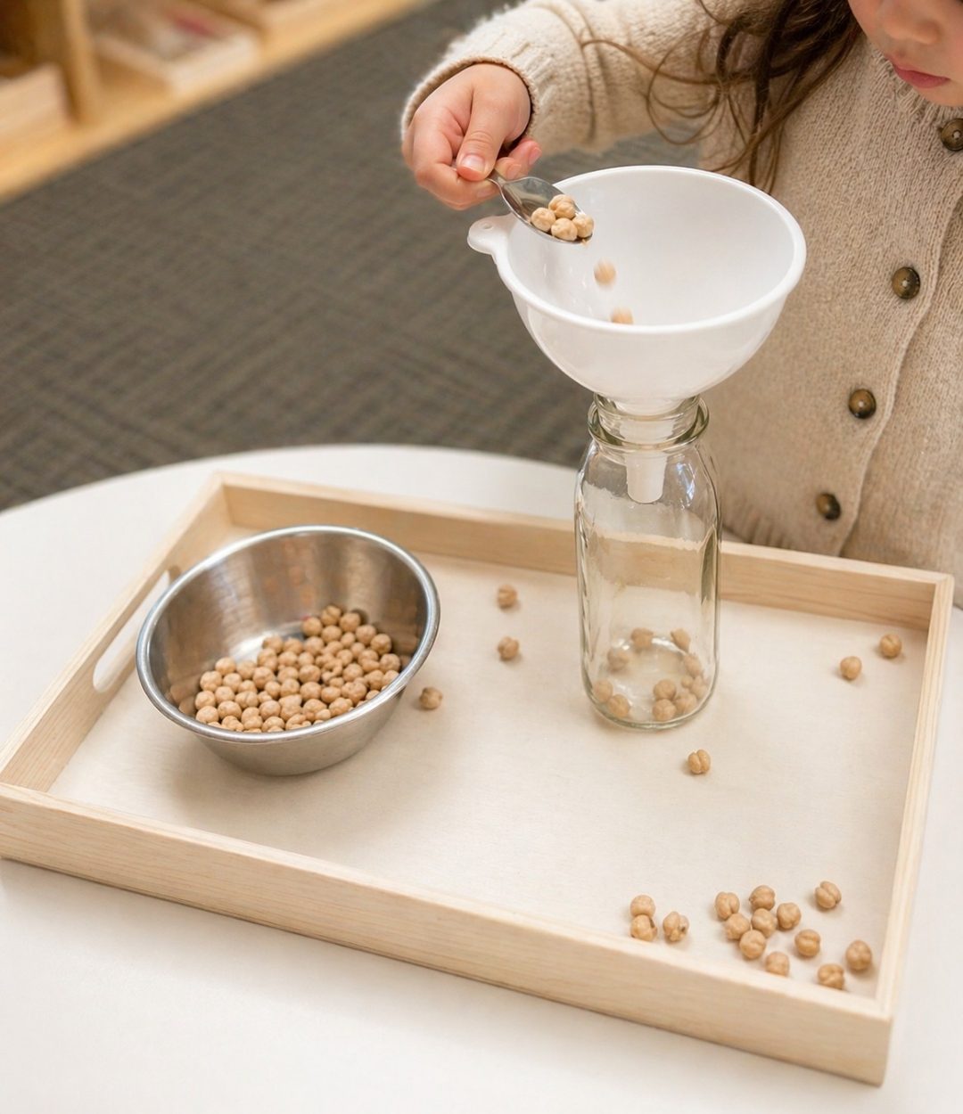 A child carefully pouring chickpeas through a funnel into a glass bottle on a Montessori practical-life tray