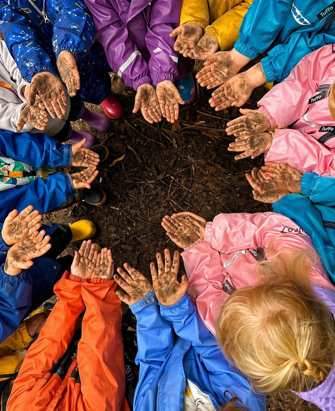 A circle of children in colourful raincoats holding out their muddy hands together after outdoor play